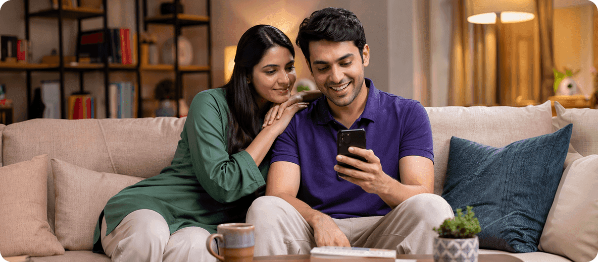 A smiling couple sits on a sofa, looking at a smartphone together.